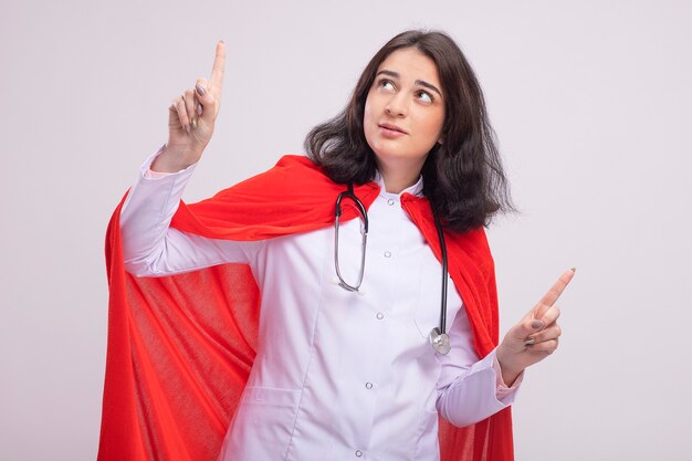 Impressionato giovane supereroe caucasica ragazza che indossa l'uniforme del medico e lo stetoscopio guardando e puntando verso l'alto