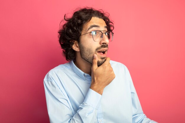 Impressionato giovane uomo bello con gli occhiali che tocca il mento guardando il lato isolato sulla parete rosa