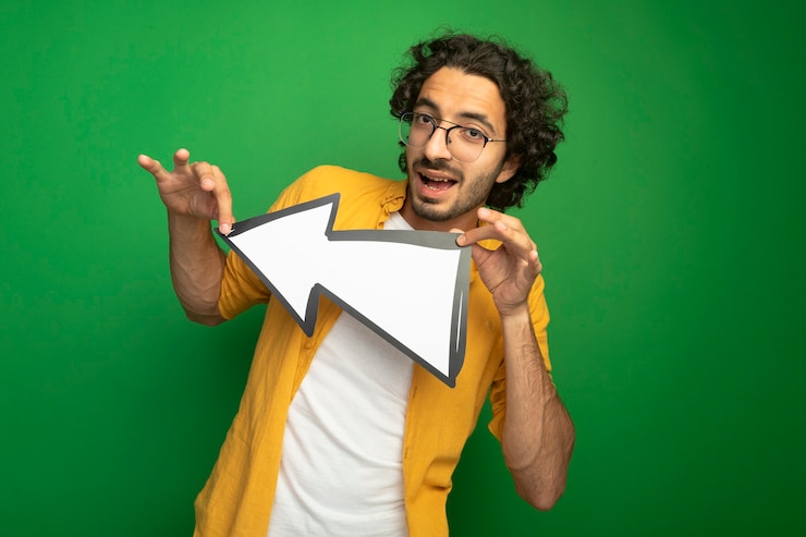 Foto gratuito impressionato giovane uomo bello con gli occhiali tenendo il segno di freccia che punta sul lato guardando la parte anteriore isolato sulla parete verde