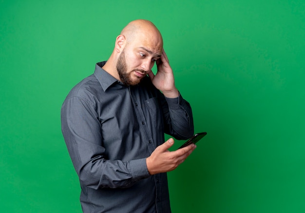 Impressionato giovane uomo calvo della call center che tiene e guardando il telefono cellulare e toccando la testa isolata sul verde con lo spazio della copia