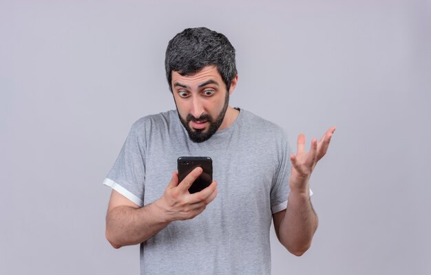 Impressionato giovane uomo caucasico bello che tiene e che esamina il telefono cellulare con la mano alzata isolata su bianco con lo spazio della copia