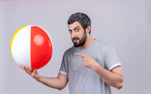 Impressionato giovane uomo caucasico bello che tiene e che punta al pallone da spiaggia isolato su bianco