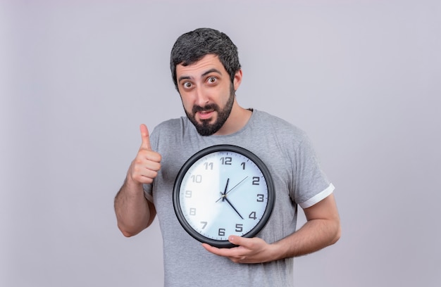 Impressionato giovane uomo caucasico bello che tiene l'orologio e che mostra il pollice su isolato su bianco con lo spazio della copia