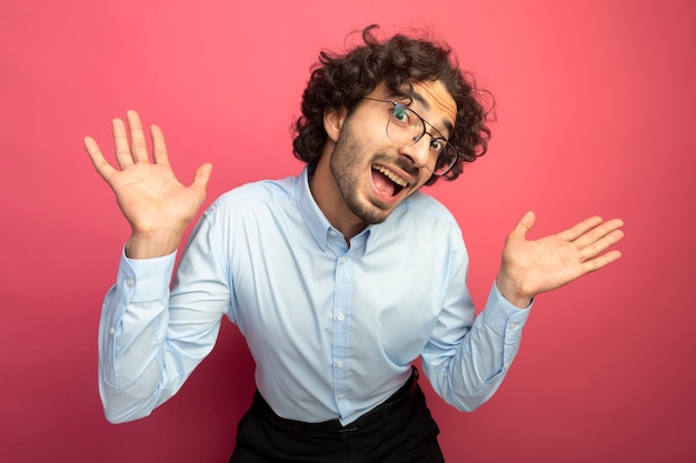 Impressionato giovane uomo caucasico bello con gli occhiali che mostra le mani vuote guardando la telecamera isolata su sfondo cremisi