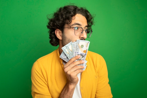 Impressionato giovane uomo caucasico bello con gli occhiali che tengono i soldi davanti alla bocca guardando il lato isolato sulla parete verde con lo spazio della copia