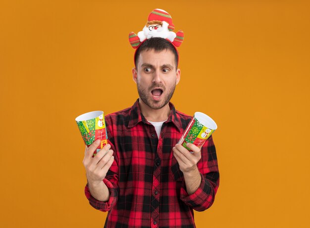 Impressionato giovane uomo caucasico che indossa la fascia di Babbo Natale che tiene i bicchieri di plastica di Natale guardando uno di loro isolato su sfondo arancione