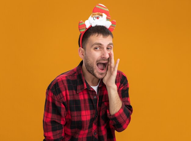 Impressionato giovane uomo caucasico che indossa la fascia di Babbo Natale tenendo la mano vicino alla bocca sussurrando isolato sulla parete arancione con lo spazio della copia