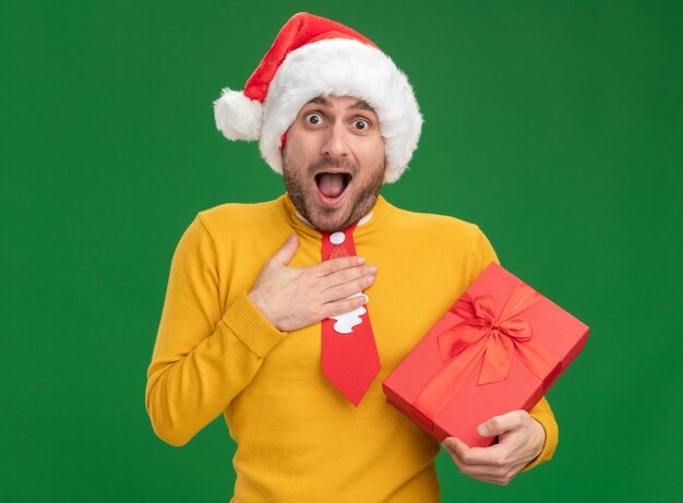 Impressionato giovane uomo caucasico indossando il cappello di Natale e cravatta tenendo il pacco regalo tenendo la mano sul petto guardando la telecamera isolata su sfondo verde