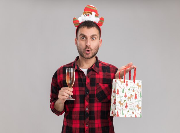 Impressionato giovane uomo caucasico indossando la fascia di Natale che tiene il sacchetto del regalo di Natale e un bicchiere di champagne guardando la telecamera isolata su sfondo bianco