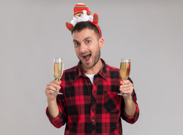 Impressionato giovane uomo caucasico indossando la fascia natalizia in possesso di due bicchieri di champagne guardando la telecamera isolata su sfondo bianco