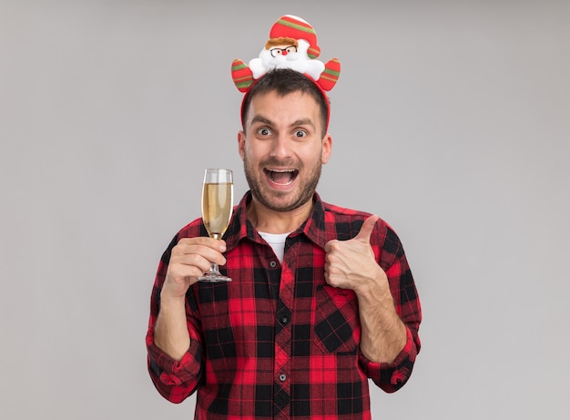 Impressionato giovane uomo caucasico indossando la fascia natalizia tenendo un bicchiere di champagne guardando la telecamera che mostra il pollice in alto isolato su sfondo bianco