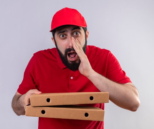Impressionato giovane uomo di consegna caucasico bello che indossa l'uniforme rossa e il cappuccio che tiene le scatole della pizza e sussurra isolato su bianco