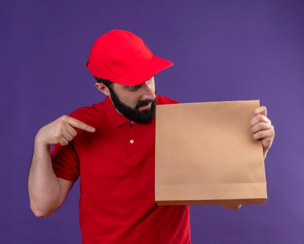 Impressionato giovane uomo di consegna caucasico bello che indossa uniforme rossa e berretto che tiene guardando e indicando il pacchetto di carta isolato sulla porpora