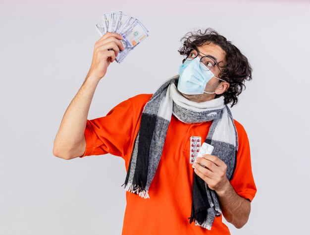 Impressionato giovane uomo malato con gli occhiali sciarpa e maschera che tiene soldi e pillole guardando soldi isolati sul muro bianco