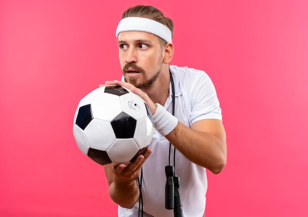 Impressionato giovane uomo sportivo bello che indossa fascia e braccialetti che tengono il pallone da calcio guardando il lato con la corda per saltare intorno al collo isolato sul muro rosa