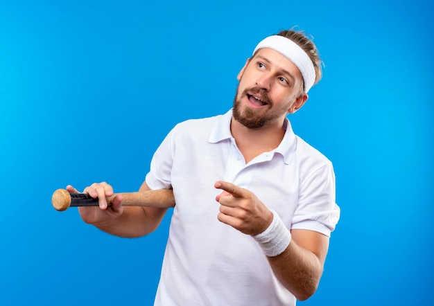 Impressionato giovane uomo sportivo bello che indossa fascia e braccialetti che tengono mazza da baseball guardando e indicando il lato isolato sulla parete blu