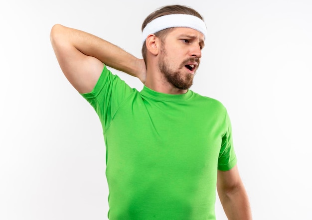 Impressionato giovane uomo sportivo bello che indossa fascia e braccialetti mettendo la mano sul collo guardando il lato isolato sul muro bianco