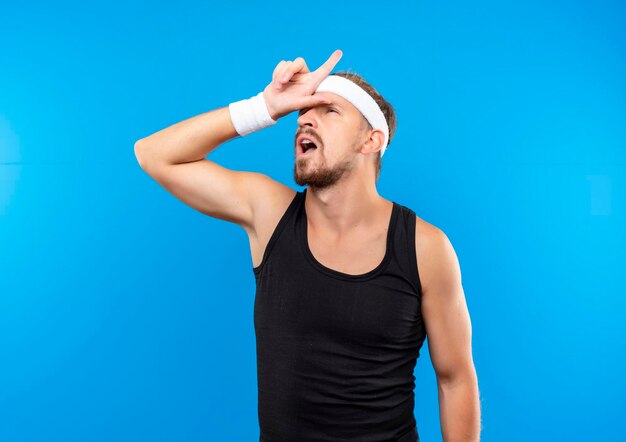 Impressionato giovane uomo sportivo bello che indossa fascia e braccialetti mettendo la mano sulla fronte e puntando verso l'alto isolato sul muro blu blue