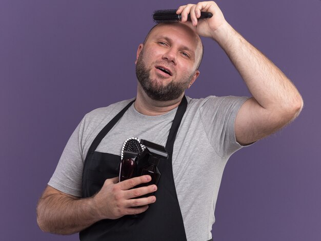 Impressionato guardando il barbiere maschio di mezza età slavo laterale in uniforme che tiene gli strumenti del barbiere e pettina i capelli isolati sul muro viola