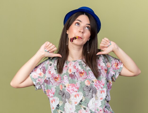 Impressionato guardando la giovane bella ragazza che indossa il cappello da festa che soffia il fischio della festa punta a se stessa