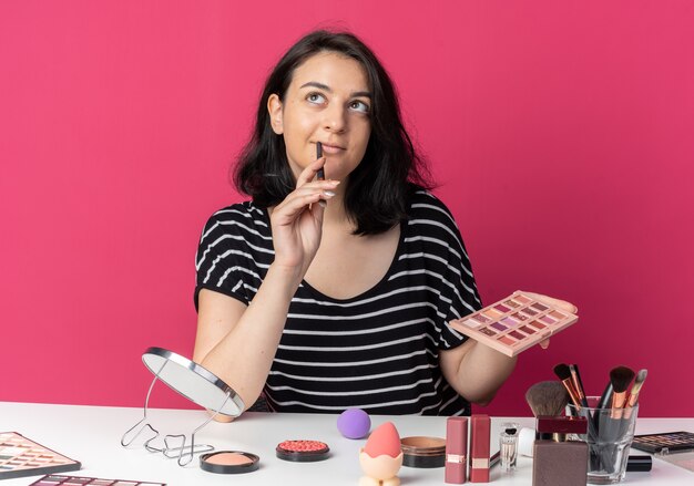 Impressionato guardando la giovane bella ragazza si siede al tavolo con gli strumenti per il trucco tenendo la tavolozza dell'ombretto con il pennello per il trucco isolato sulla parete rosa