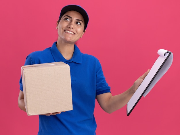 Impressionato guardando la giovane ragazza di consegna laterale che indossa l'uniforme con cappuccio che tiene la scatola con appunti isolato sulla parete rosa