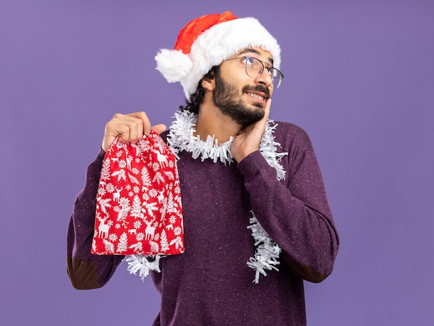 Impressionato guardando lato giovane bel ragazzo che indossa il cappello di natale con la ghirlanda sul collo che tiene la borsa di natale mettendo la mano sulla guancia isolata sulla parete blu