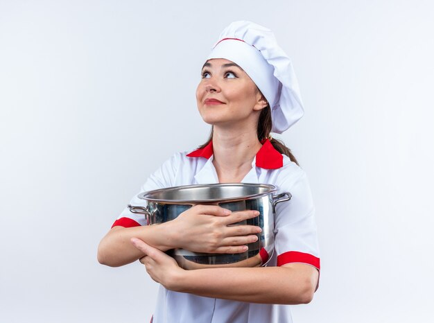 impressionato guardando lato giovane cuoca che indossa l'uniforme dello chef tenendo la casseruola isolata sul muro bianco