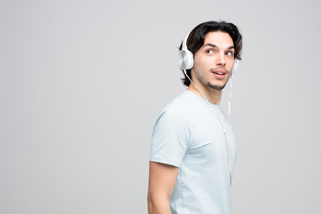 impressionato il giovane bell'uomo che indossa le cuffie in piedi nella vista di profilo guardando il lato isolato su sfondo bianco con spazio per la copia