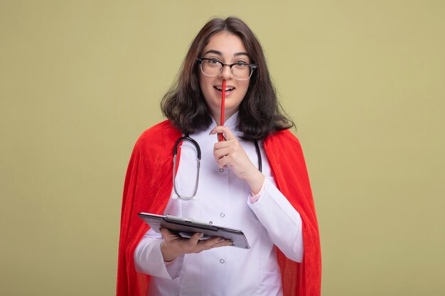 Impressionato la ragazza del supereroe in mantello rosso che indossa l'uniforme del medico e uno stetoscopio con gli occhiali che tengono gli appunti guardando davanti toccando le labbra con una matita