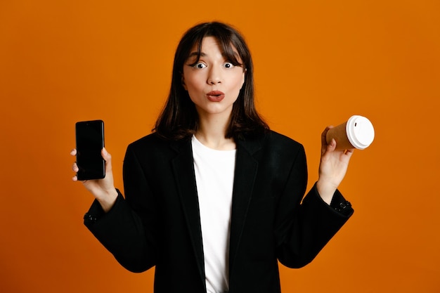 Impressionato tenendo la tazza di caffè con il telefono giovane bella donna che indossa una giacca nera isolata su sfondo arancione