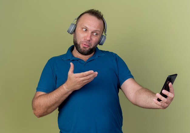 Impressionato uomo adulto slavo che indossa le cuffie tenendo guardando e indicando con la mano al telefono cellulare