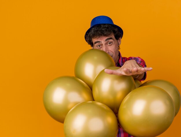 Impressionato uomo caucasico di mezza età del partito che indossa il cappello del partito in piedi dietro i palloncini che allunga la mano verso isolato sulla parete arancione con lo spazio della copia