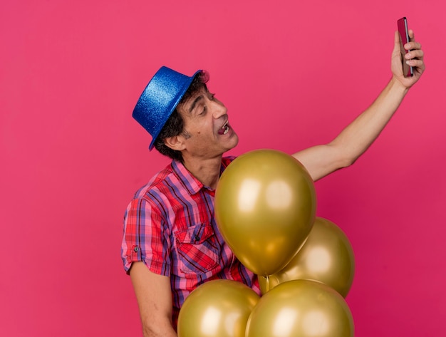 Impressionato uomo caucasico di mezza età del partito che indossa il cappello del partito in piedi dietro i palloncini che prendono selfie isolato su priorità bassa cremisi con lo spazio della copia