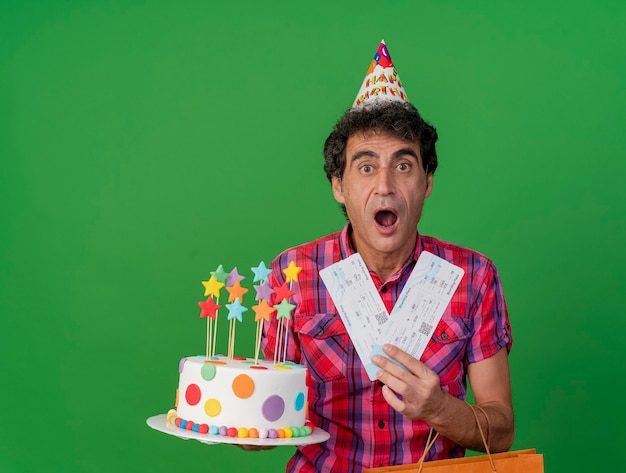 Impressionato uomo caucasico di mezza età del partito che indossa il cappello di compleanno che tiene il sacchetto di carta della torta di compleanno e biglietti aerei che guarda l'obbiettivo isolato su priorità bassa verde con lo spazio della copia