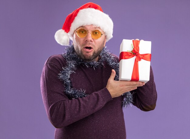 Impressionato uomo di mezza età che indossa un cappello da Babbo Natale e una ghirlanda di orpelli intorno al collo con gli occhiali che tiene il pacchetto regalo guardando la telecamera isolata su sfondo viola