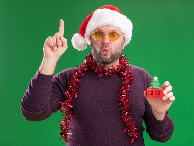 Impressionato uomo di mezza età che indossa un cappello di Babbo Natale e una ghirlanda di orpelli intorno al collo con gli occhiali che tengono il giocattolo dell'albero di Natale con la data rivolta verso l'alto con le labbra increspate isolate sul muro verde