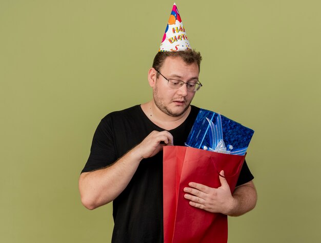 Impressionato uomo slavo adulto in occhiali ottici che indossa un cappello di compleanno tiene e guarda la confezione regalo in una borsa della spesa di carta