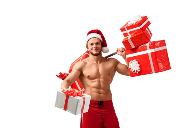 In esclusiva per te. Uomo tonico in palestra a torso nudo che indossa il cappello di Babbo Natale che trasporta pesi con regali che porge un regalo guardando allegramente e sorridendo alla telecamera, 2018, 2019.