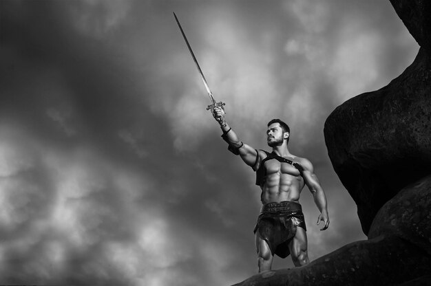 In nome di Dio. Ritratto monocromatico di un potente gladiatore muscolare che tiene la spada fino al cielo tempestoso copyspace