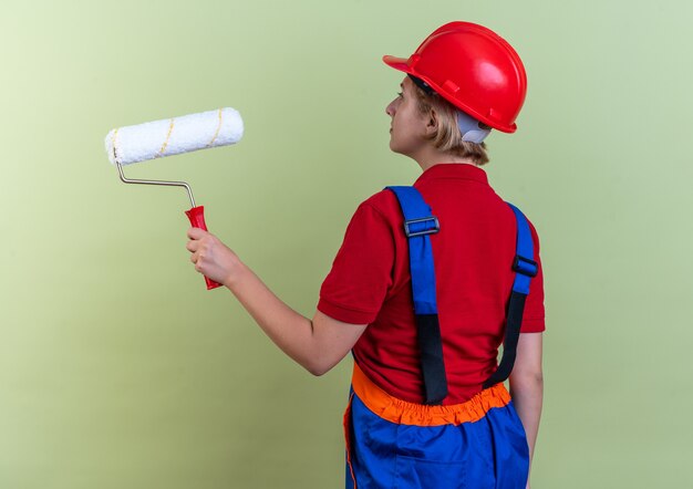 in piedi dietro la vista giovane donna del costruttore in uniforme che tiene la spazzola a rullo isolata sulla parete verde oliva con spazio di copia