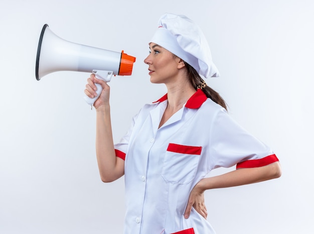 In piedi in vista di profilo giovane cuoca che indossa l'uniforme da chef parla sull'altoparlante mettendo la mano sull'anca isolata sul muro bianco white