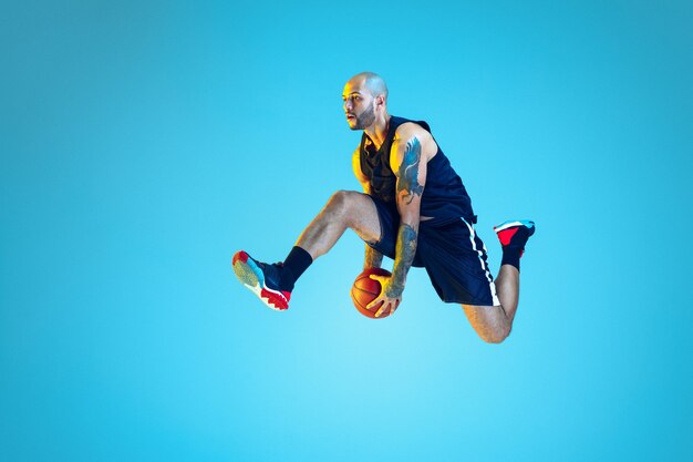 In salto. Giovane giocatore di basket della squadra che indossa la formazione di abbigliamento sportivo, pratica in azione, movimento sulla parete blu in luce al neon. Concetto di sport, movimento, energia e stile di vita dinamico e sano.
