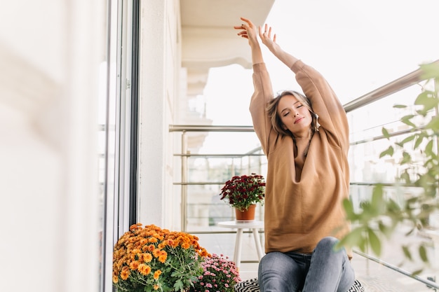 Incantevole giovane donna che si estende al balcone. Ritratto dell'interno della ragazza bionda soddisfatta che posa con le mani in alto.