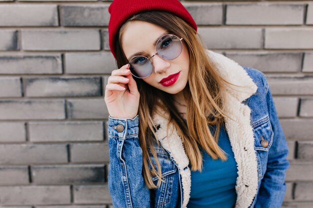 Incantevole giovane signora in occhiali rotondi blu in posa davanti al muro di mattoni. Colpo esterno di gioiosa ragazza caucasica con la pelle bianca indossa giacca di jeans.