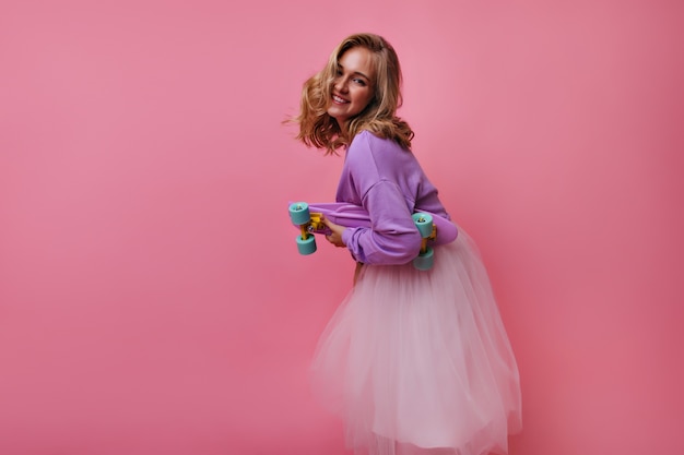 Incantevole modello femminile in gonna lussureggiante e camicia viola in posa con lo skateboard. Ritratto dell'interno della ragazza bionda interessata scherzosamente