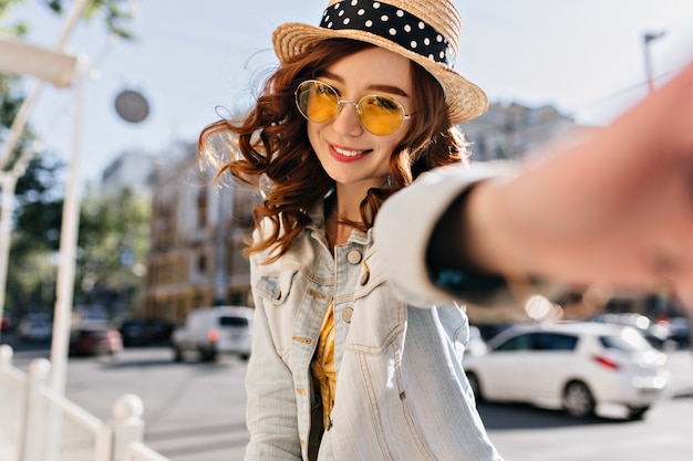 Incantevole ragazza caucasica con capelli ricci rossi che fa selfie sulla strada. Giovane signora allegra in giacca di jeans che ride sulla città.