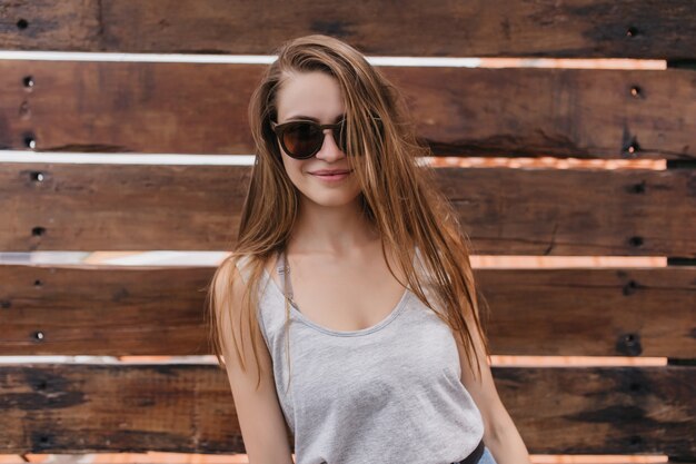 Incantevole ragazza con pelle bianca in piedi sulla parete in legno. Foto di felice giovane donna in occhiali da sole in posa nella calda giornata di primavera con un sorriso romantico.