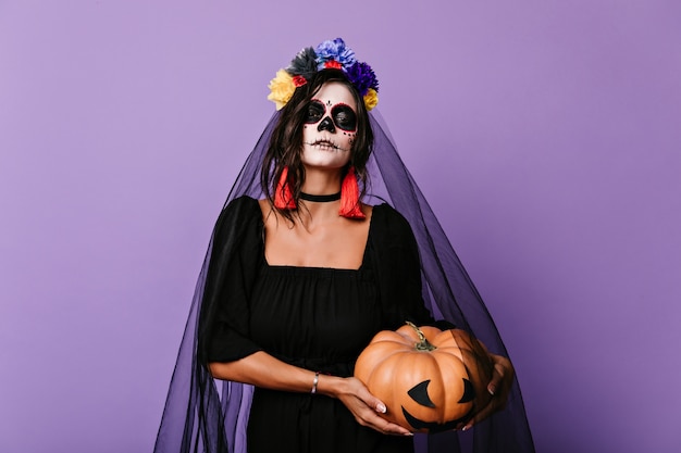 Incantevole ragazza con trucco messicano spaventoso che prepara per halloween. Tiro al coperto di romantica sposa morta in velo nero che tiene la zucca.