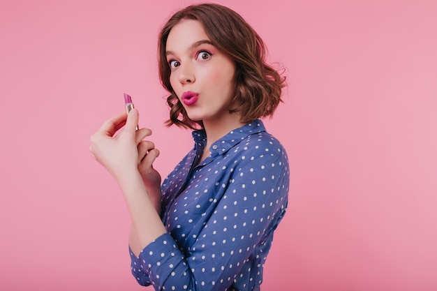 Incantevole ragazza dagli occhi marroni con i capelli corti ricci che scherza mentre fa il trucco. Ritratto dell'interno del modello femminile allegro in camicetta che posa con il rossetto sulla parete rosa.